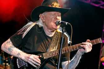 Johnny Winter performing on stage with a guitar, wearing a hat and black shirt.