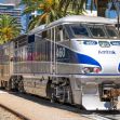 An Amtrak train stopped at a station surrounded by palm trees and buildings. An Amtrak train stopped at a station surrounded by palm trees and buildings.
