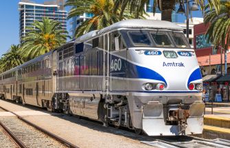 An Amtrak train stopped at a station surrounded by palm trees and buildings.