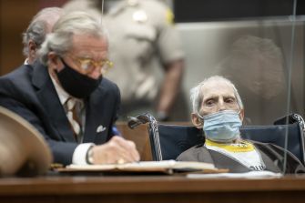 Robert Durst sitting in a courtroom with his lawyer, wearing a mask and looking frail, surrounded by law enforcement officers.