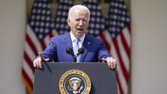 President Biden addressing the public at a podium with the Presidential seal, flanked by decorative American flags in the background.