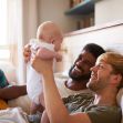 Two smiling men playing with a baby while lying down on a bed, showing a warm and loving family moment. Two smiling men playing with a baby while lying down on a bed, showing a warm and loving family moment.