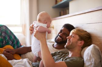 Two smiling men playing with a baby while lying down on a bed, showing a warm and loving family moment.