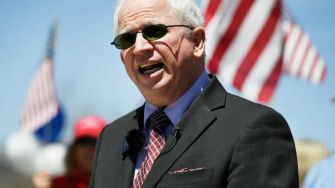 John Eastman speaking at a rally, with American flags visible in the background.