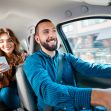 A rideshare driver smiling while driving with a passenger in a car. A rideshare driver smiling while driving with a passenger in a car.