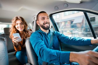 A rideshare driver smiling while driving with a passenger in a car.