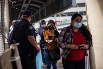 People walking through a border checkpoint, some wearing masks, with a uniformed officer present.