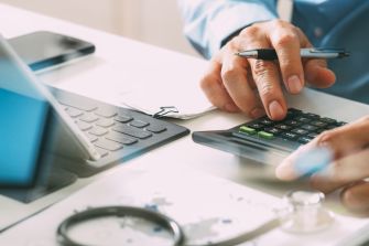 A person using a calculator and writing notes at a desk with a laptop and medical equipment.