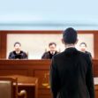 A defendant standing in a courtroom facing three judges. A defendant standing in a courtroom facing three judges.