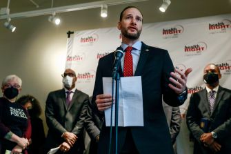 A man speaking at a press conference, holding papers, with attendees in the background wearing face masks.