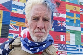 A man with a patriotic outfit and facial expression standing in front of a wall of international flags.