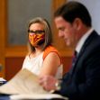 A woman wearing a mask with the Arizona flag design and a man in a suit are seated at a table during a discussion or press conference. A woman wearing a mask with the Arizona flag design and a man in a suit are seated at a table during a discussion or press conference.