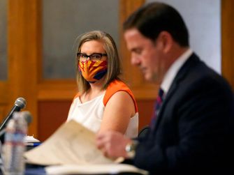 A woman wearing a mask with the Arizona flag design and a man in a suit are seated at a table during a discussion or press conference.