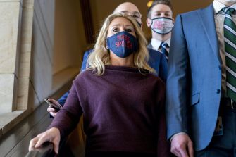 Marjorie Taylor Greene walking down a staircase, wearing a mask that says "FREE SPEECH," accompanied by several individuals in suits.