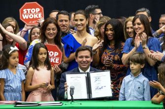 A group of people, including children, gathers around Florida Governor Ron DeSantis as he holds a signed document related to the "Stop WOKE Act," with signs displaying the phrase "STOP WOKE" in the background.