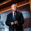 Sen. Lindsey Graham standing in front of a backdrop featuring the U.S. Capitol and an American flag. Sen. Lindsey Graham standing in front of a backdrop featuring the U.S. Capitol and an American flag.