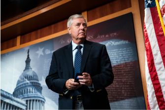 Sen. Lindsey Graham standing in front of a backdrop featuring the U.S. Capitol and an American flag.