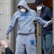 A man in a gray "REBEL" hoodie and sweatpants exits a building, accompanied by another man, while wearing masks. A man in a gray "REBEL" hoodie and sweatpants exits a building, accompanied by another man, while wearing masks.