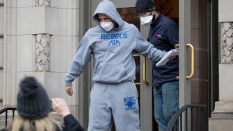 A man in a gray "REBEL" hoodie and sweatpants exits a building, accompanied by another man, while wearing masks.