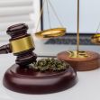 A gavel resting on a wooden base alongside scales of justice and marijuana on a desk with a laptop in the background. A gavel resting on a wooden base alongside scales of justice and marijuana on a desk with a laptop in the background.