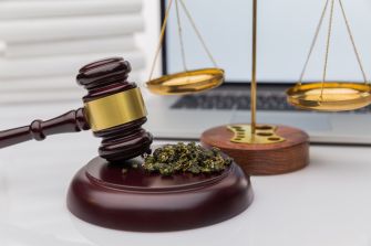 A gavel resting on a wooden base alongside scales of justice and marijuana on a desk with a laptop in the background.