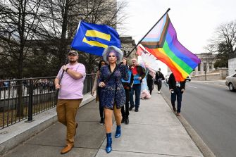 A diverse group of people, including a drag performer, marching with LGBTQ+ flags in a demonstration.