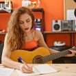 A woman with curly hair playing an acoustic guitar while writing in a notebook in a music studio. A woman with curly hair playing an acoustic guitar while writing in a notebook in a music studio.