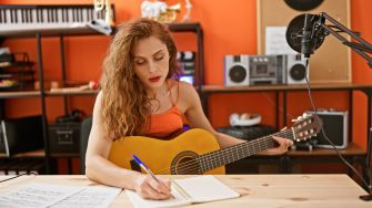 A woman with curly hair playing an acoustic guitar while writing in a notebook in a music studio.