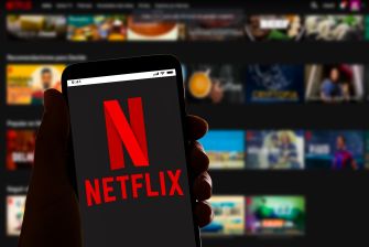 A person holding a smartphone displaying the Netflix logo, with a blurred background of Netflix content.