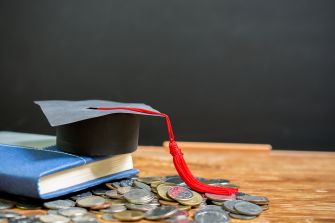 A graduation cap resting on a stack of coins next to a book, symbolizing student debt and education finances.