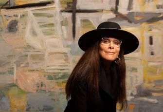 A woman wearing a black hat and glasses poses in front of an abstract background.