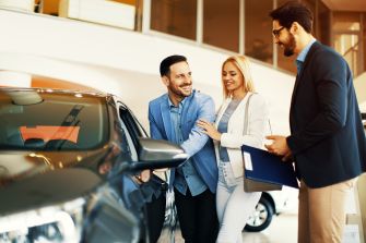 A couple interacting with a car salesman at a dealership, looking at a vehicle.