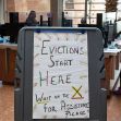Sign indicating the start of evictions and requesting individuals to wait for assistance, with people working in the background. Sign indicating the start of evictions and requesting individuals to wait for assistance, with people working in the background.