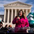 A woman in a pink coat addresses a crowd with balloons in front of the U.S. Supreme Court building. A woman in a pink coat addresses a crowd with balloons in front of the U.S. Supreme Court building.