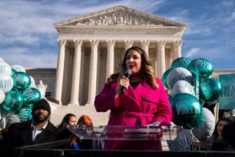 A woman in a pink coat addresses a crowd with balloons in front of the U.S. Supreme Court building.