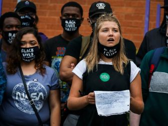 A group of individuals wearing masks with the Black Lives Matter slogan at a protest or gathering. One person is speaking while holding a piece of paper.