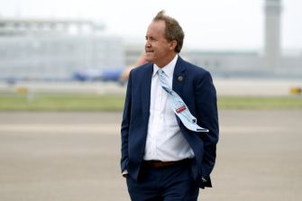 A man in a suit walking on an airport tarmac, appearing contemplative.