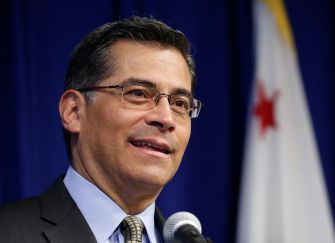 A man in glasses speaking at a microphone with a California flag in the background.