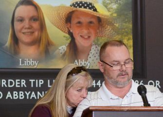 A man speaking at a press conference with images of two teens, Libby German and Abby Williams, displayed in the background.