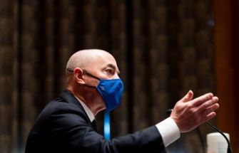 A man in a suit gesturing while speaking, wearing a blue face mask, during a congressional hearing.