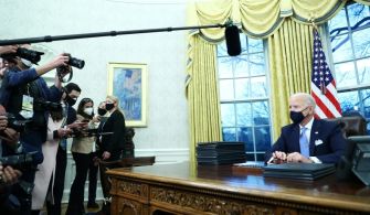 President Biden seated at the Resolute Desk in the Oval Office surrounded by reporters on the day he signed multiple executive orders.