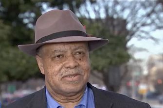 A man wearing a hat speaks outdoors in front of blurred trees.