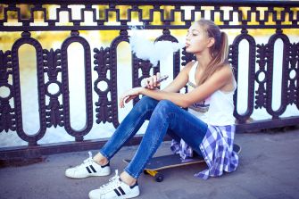 A young person sitting on a skateboard by a waterfront, exhaling vapor from an e-cigarette.