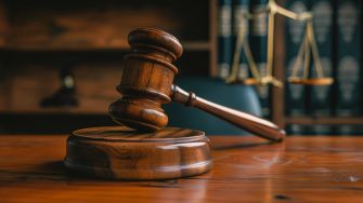 Gavel on a wooden court table with a backdrop of legal books and scales of justice.
