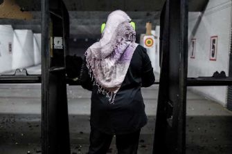 A woman wearing a hijab stands at a gun range, facing away from the camera, with shooting targets visible in the background.