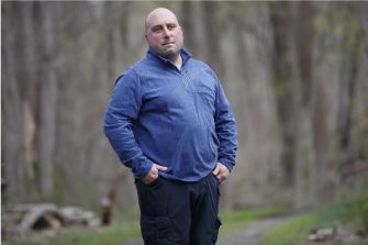 A man standing in a wooded area, wearing a blue jacket and looking directly at the camera.