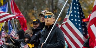 A group of people at a rally holding flags, with one person wearing a mask resembling a former president.