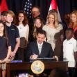 A group of people, including children, surrounds a man signing a document at a table, with flags in the background. A group of people, including children, surrounds a man signing a document at a table, with flags in the background.