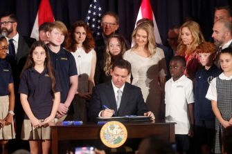 A group of people, including children, surrounds a man signing a document at a table, with flags in the background.