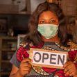 A woman wearing a mask stands in a doorway holding an "OPEN" sign. A woman wearing a mask stands in a doorway holding an "OPEN" sign.